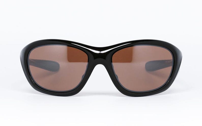K&ouml;nnte beinhalten: Schwarze Sonnenbrille mit braunen Gl&auml;sern. Die Sonnenbrille hat einen dicken, abgerundeten Rahmen und ein geschwungenes Design. Die Gl&auml;ser sind warmbraun und der Rahmen ist gl&auml;nzend schwarz. Die Sonnenbrille ist vor einem wei&szlig;en Hintergrund platziert.