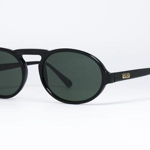 May include: Black sunglasses with dark green lenses. The sunglasses have a rounded shape with a bridge connecting the two lenses. The arms of the sunglasses are straight and have a gold logo on the side.