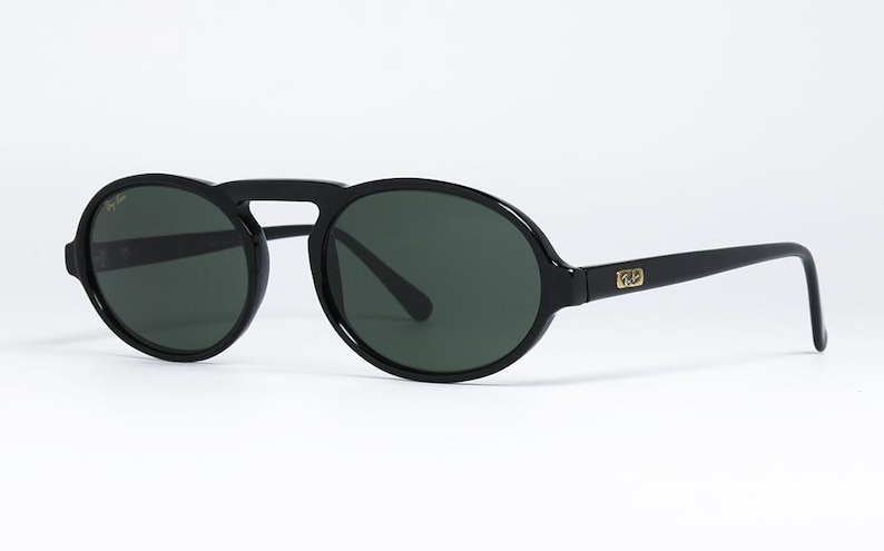 May include: Black sunglasses with dark green lenses. The sunglasses have a rounded shape with a bridge connecting the two lenses. The arms of the sunglasses are straight and have a gold logo on the side.