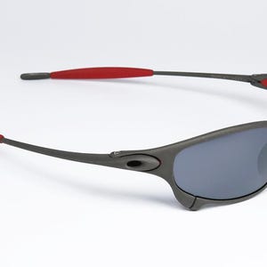 May include: Dark gray sunglasses with gray lenses and red accents on the temples. The sunglasses have a sporty, wraparound design. The frame is a matte finish, and the lenses are a dark gray color.