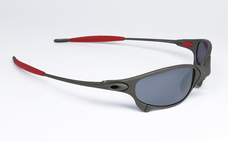 May include: Dark gray sunglasses with gray lenses and red accents on the temples. The sunglasses have a sporty, wraparound design. The frame is a matte finish, and the lenses are a dark gray color.