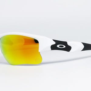May include: White and black sunglasses with orange and yellow lenses. The sunglasses have a sleek design with a black and white pattern on the arms and a small blue accent. The lenses have a mirrored finish.