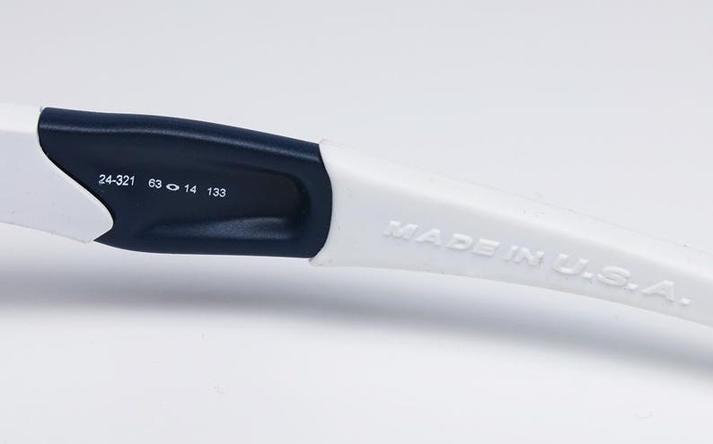 以下が含まれることがあります： 白とネイビーブルーのメガネアームのクローズアップ。テキスト「MADE IN U.S.A.」は白いアームにエンボス加工されています。アームには「24-321 63 14 133」の数字も印刷されています。