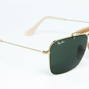 May include: Gold-framed sunglasses with dark green lenses. The sunglasses feature a double bridge, a light brown brow bar, and light brown temples. The Ray-Ban logo is visible on the lens.