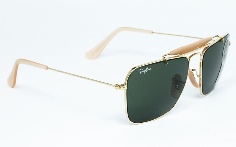 May include: Gold-framed sunglasses with dark green lenses. The sunglasses feature a double bridge, a light brown brow bar, and light brown temples. The Ray-Ban logo is visible on the lens.