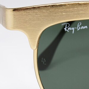Puede incluir: Primer plano de gafas de sol Ray-Ban con montura dorada y lentes verdes. El logotipo de Ray-Ban es visible en la lente. La montura tiene un acabado texturizado. Fondo blanco.