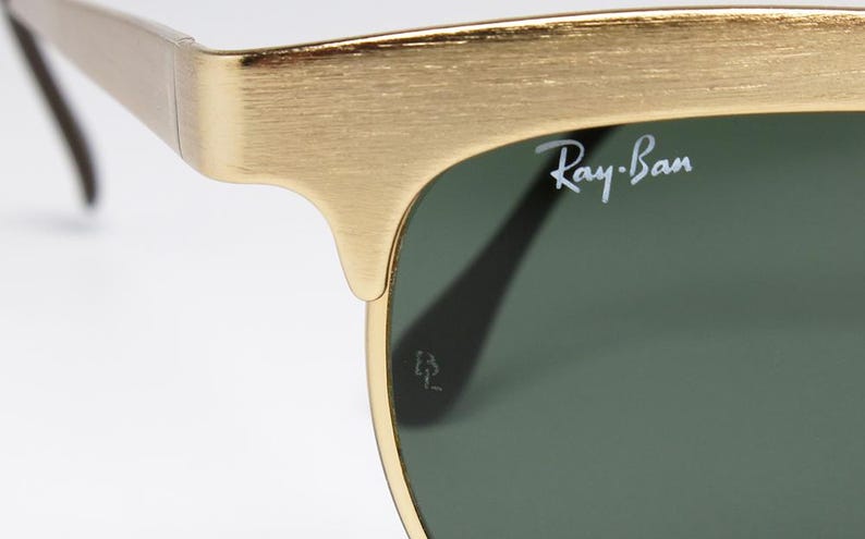 Puede incluir: Primer plano de gafas de sol Ray-Ban con montura dorada y lentes verdes. El logotipo de Ray-Ban es visible en la lente. La montura tiene un acabado texturizado. Fondo blanco.