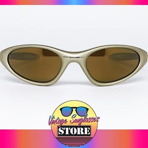 May include: Gold-tone Oakley sunglasses with brown lenses. The sunglasses have a curved frame and the Oakley logo is visible on the bridge. The image includes a logo for Vintage Sunglasses Store.