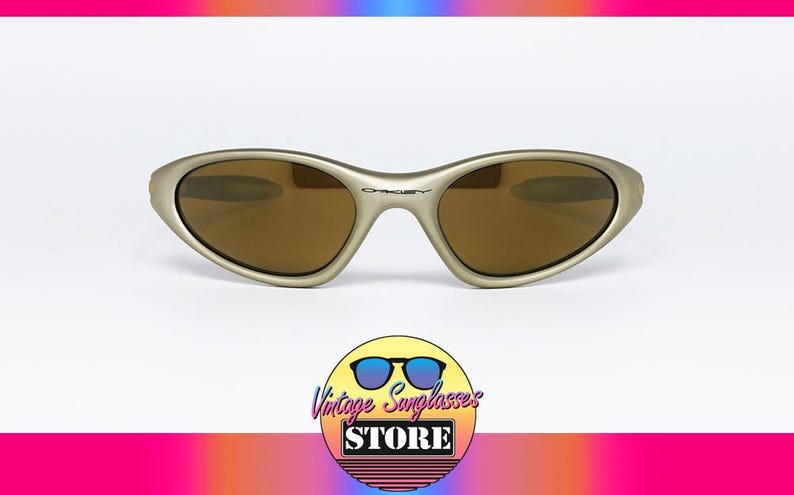 May include: Gold-tone Oakley sunglasses with brown lenses. The sunglasses have a curved frame and the Oakley logo is visible on the bridge. The image includes a logo for Vintage Sunglasses Store.