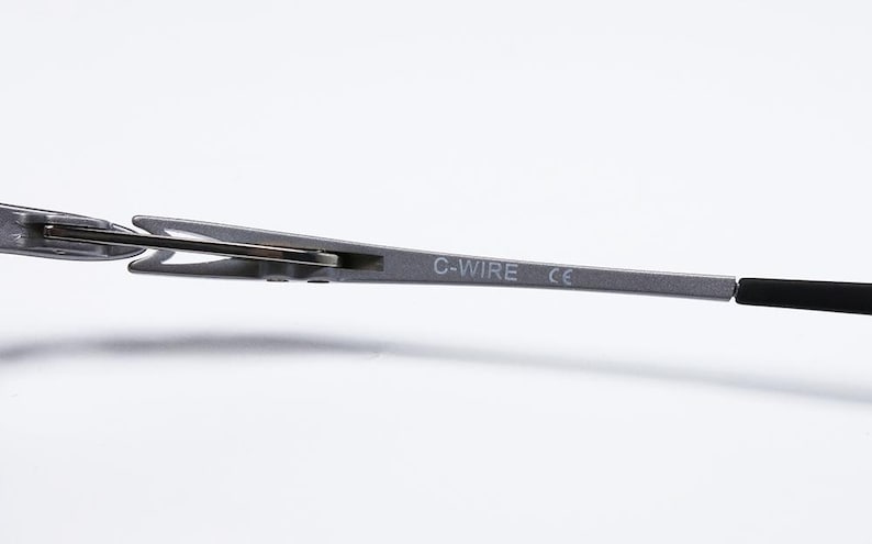 May include: Close-up of a silver metal eyeglass frame arm with the text "C-WIRE" and "CE" printed on it. The arm connects to a black temple tip. The frame is set against a white background.