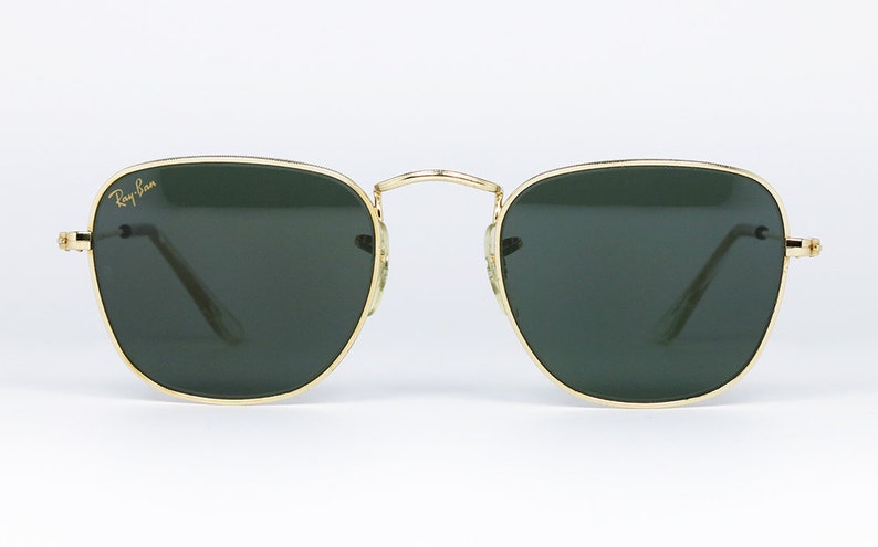 May include: Square sunglasses with gold frames and green lenses. The sunglasses feature the Ray-Ban logo on the left lens.