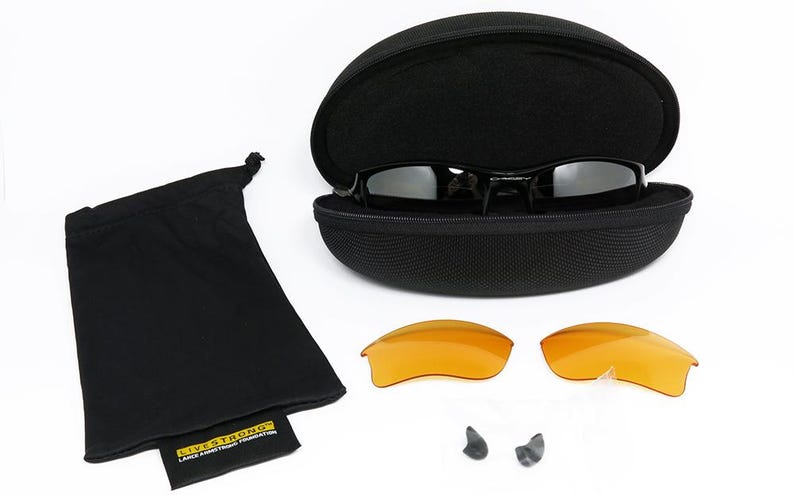 K&ouml;nnte beinhalten: Schwarze Sonnenbrille mit schwarzem Etui, zwei orangefarbenen Gl&auml;sern, einem schwarzen Kordelzugbeutel und zwei Nasenpads. Die Sonnenbrille befindet sich in einem offenen schwarzen Etui. Der Kordelzugbeutel hat ein kleines Etikett mit der Aufschrift "LIVE STRONG".