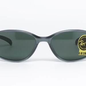 May include: Dark gray Ray-Ban sunglasses with oval lenses. The sunglasses have a black frame and a yellow sticker with the Ray-Ban logo and "G-15 LENS" text. The Ray-Ban logo is also on the left arm.