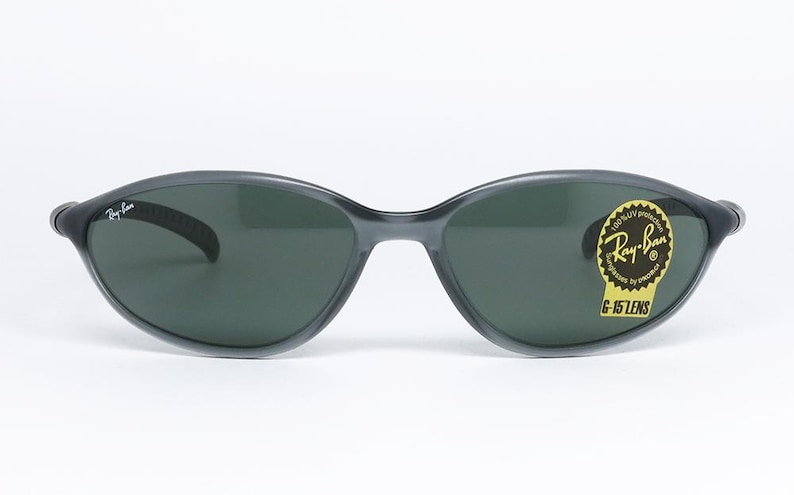 May include: Dark gray Ray-Ban sunglasses with oval lenses. The sunglasses have a black frame and a yellow sticker with the Ray-Ban logo and "G-15 LENS" text. The Ray-Ban logo is also on the left arm.