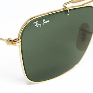 May include: Close-up of Ray-Ban sunglasses with a gold metal frame and dark green lenses. The Ray-Ban logo is visible on the lens. The sunglasses have a classic aviator design with a double bridge and nose pads.