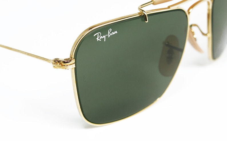 May include: Close-up of Ray-Ban sunglasses with a gold metal frame and dark green lenses. The Ray-Ban logo is visible on the lens. The sunglasses have a classic aviator design with a double bridge and nose pads.