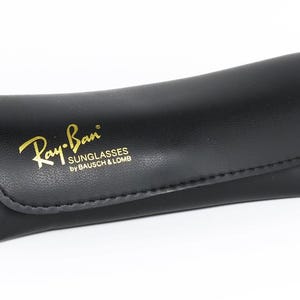 Pu&ograve; includere: Custodia per occhiali da sole in pelle nera con il logo Ray-Ban in lettere dorate. La custodia &egrave; curva e ha una chiusura a patta. Il testo "SUNGLASSES by BAUSCH & LOMB" &egrave; sotto il logo.