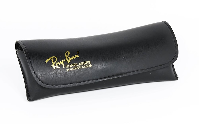 Pu&ograve; includere: Custodia per occhiali da sole in pelle nera con il logo Ray-Ban in lettere dorate. La custodia &egrave; curva e ha una chiusura a patta. Il testo "SUNGLASSES by BAUSCH & LOMB" &egrave; sotto il logo.