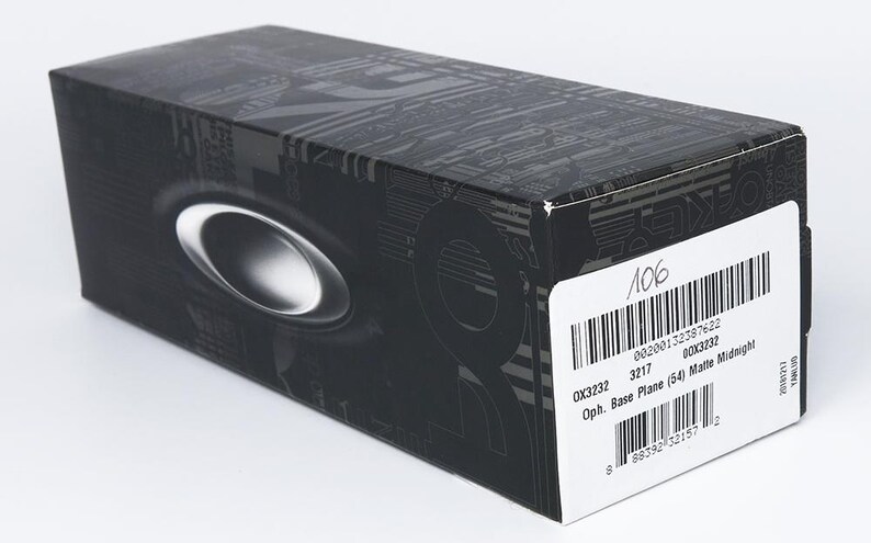 K&ouml;nnte beinhalten: Eine schwarze rechteckige Box mit einem silbernen ovalen Emblem. Die Box hat ein gemustertes Design und ein wei&szlig;es Etikett mit Text und einem Barcode. Das Etikett enth&auml;lt die Texte "OX3232" und "Base Plane (54) Matte Midnight".