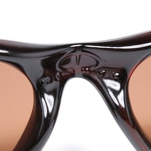 Pode incluir: Close-up de &oacute;culos de sol castanhos com arma&ccedil;&atilde;o escura. As lentes s&atilde;o de cor castanho claro. A ponte dos &oacute;culos de sol apresenta um pequeno emblema. Os &oacute;culos de sol s&atilde;o de estilo cl&aacute;ssico.