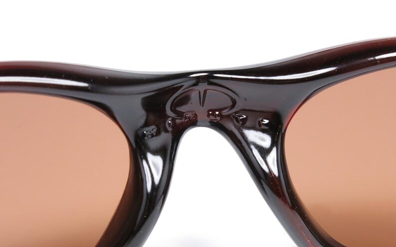 Pode incluir: Close-up de &oacute;culos de sol castanhos com arma&ccedil;&atilde;o escura. As lentes s&atilde;o de cor castanho claro. A ponte dos &oacute;culos de sol apresenta um pequeno emblema. Os &oacute;culos de sol s&atilde;o de estilo cl&aacute;ssico.