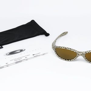 May include: Sunglasses with a leopard print frame and brown lenses. The image also shows a black sunglasses case and a white information card. The Oakley logo is visible on the case and card.