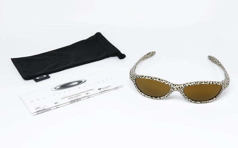 May include: Sunglasses with a leopard print frame and brown lenses. The image also shows a black sunglasses case and a white information card. The Oakley logo is visible on the case and card.