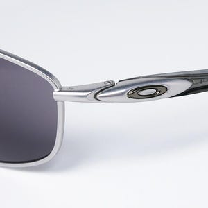 May include: Close-up of a pair of Oakley sunglasses. The sunglasses have a silver frame and dark gray lenses. The arm of the glasses features the Oakley logo in black and silver.