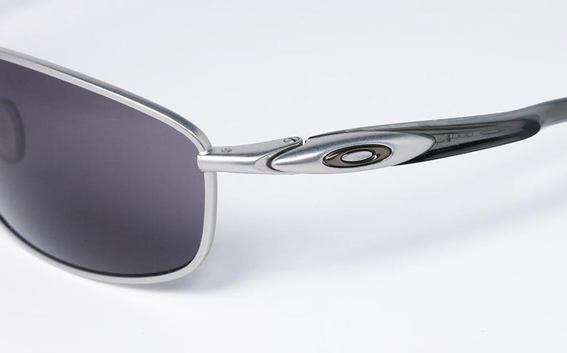 May include: Close-up of a pair of Oakley sunglasses. The sunglasses have a silver frame and dark gray lenses. The arm of the glasses features the Oakley logo in black and silver.