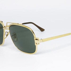Puede incluir: Gafas de sol con montura dorada y lentes verde oscuro. Las gafas tienen una forma de aviador cl&aacute;sica con doble puente y montura de metal dorado. Las patillas son doradas con puntas marrones. Sobre fondo blanco.