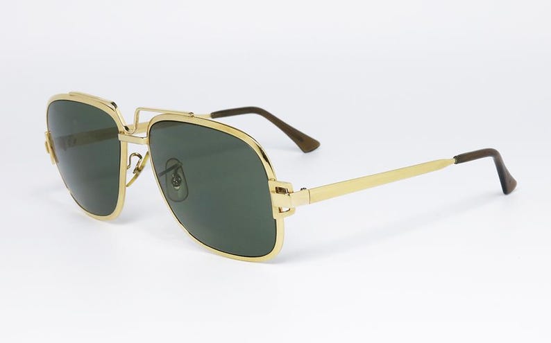 Puede incluir: Gafas de sol con montura dorada y lentes verde oscuro. Las gafas tienen una forma de aviador cl&aacute;sica con doble puente y montura de metal dorado. Las patillas son doradas con puntas marrones. Sobre fondo blanco.