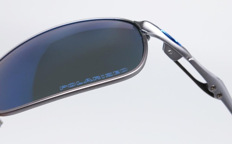 May include: Close-up of polarized sunglasses with a silver frame and blue lenses. The word "POLARIZED" is printed in blue on the lens. The sunglasses have a modern, sporty design.