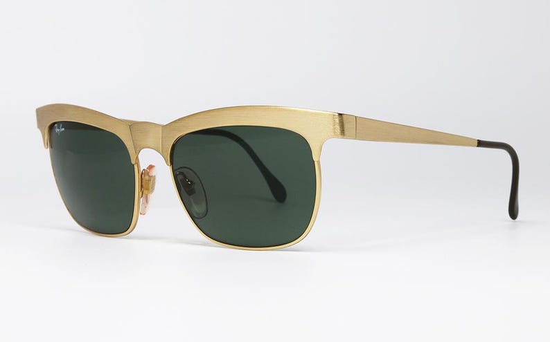 Puede incluir: Gafas de sol doradas con lentes verde oscuro. Las monturas tienen un dise&ntilde;o distintivo de cejas y patillas elegantes. Las lentes son de color verde oscuro y las gafas de sol tienen un estilo cl&aacute;sico y atemporal.