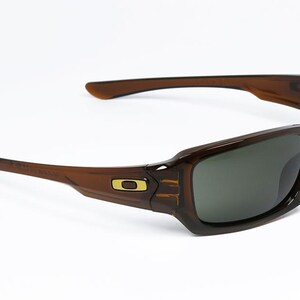 May include: Brown sunglasses with a rectangular frame and dark lenses. The sunglasses have a curved design with a gold logo on the side. The arms are a lighter brown colour.