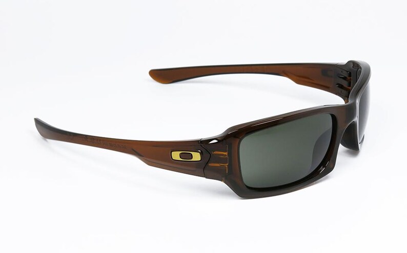 May include: Brown sunglasses with a rectangular frame and dark lenses. The sunglasses have a curved design with a gold logo on the side. The arms are a lighter brown colour.