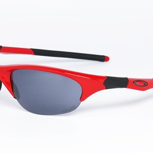 Puede incluir: Gafas de sol rojas y negras con lentes grises. Las gafas tienen un dise&ntilde;o deportivo con montura roja y detalles negros en las patillas. El logotipo de Oakley es visible en la patilla.