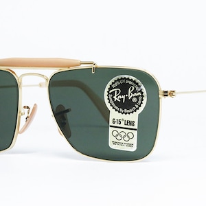 May include: Gold-framed Ray-Ban sunglasses with green lenses. The sunglasses feature a light brown bridge and temple tips. The lenses have a sticker that reads "Ray-Ban" and "G-15 LENS".