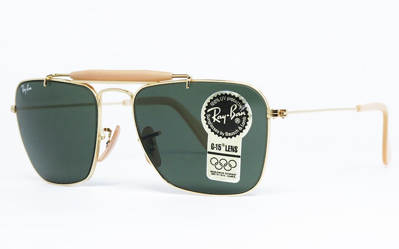 May include: Gold-framed Ray-Ban sunglasses with green lenses. The sunglasses feature a light brown bridge and temple tips. The lenses have a sticker that reads "Ray-Ban" and "G-15 LENS".