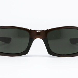 May include: Brown sunglasses with dark lenses. The sunglasses have a rectangular shape with rounded edges. The frame is a solid brown colour. The lenses are dark grey, providing protection from the sun.
