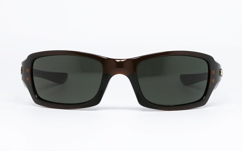 May include: Brown sunglasses with dark lenses. The sunglasses have a rectangular shape with rounded edges. The frame is a solid brown colour. The lenses are dark grey, providing protection from the sun.
