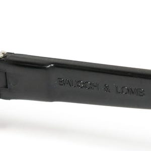 Op de afbeelding: Close-up van een zwarte zonnebrilarm met de tekst "BAUSCH & LOMB" in reli&euml;f aan de zijkant. De lens heeft een warme bruine kleur. Het scharnier is zichtbaar met een kleine zilveren schroef.
