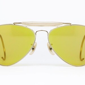 K&ouml;nnte beinhalten: Goldene Aviator-Sonnenbrille mit gelben Gl&auml;sern. Die Sonnenbrille hat einen Metallrahmen und eine Br&uuml;cke, die die beiden Gl&auml;ser verbindet.