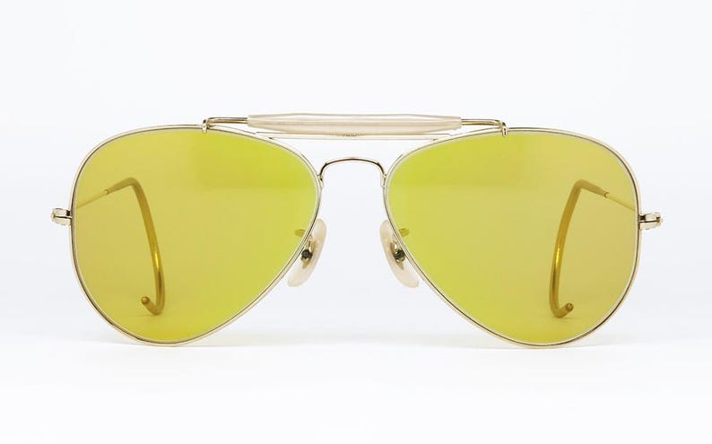 K&ouml;nnte beinhalten: Goldene Aviator-Sonnenbrille mit gelben Gl&auml;sern. Die Sonnenbrille hat einen Metallrahmen und eine Br&uuml;cke, die die beiden Gl&auml;ser verbindet.