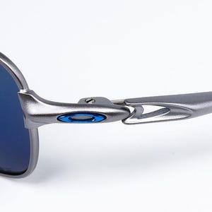 May include: Close-up of a pair of sunglasses with a metallic gray frame and blue lenses. The frame features a blue accent near the hinge. The sunglasses are designed for outdoor use and offer protection from the sun.