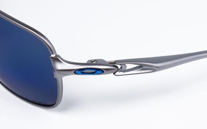 May include: Close-up of a pair of sunglasses with a metallic gray frame and blue lenses. The frame features a blue accent near the hinge. The sunglasses are designed for outdoor use and offer protection from the sun.