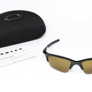 May include: Black sunglasses with brown lenses and a black case. The sunglasses have a sleek design with a curved frame. A warranty card and the case are also visible. The brand name "OAKLEY" is printed on the warranty card.