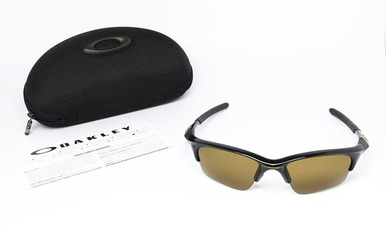 May include: Black sunglasses with brown lenses and a black case. The sunglasses have a sleek design with a curved frame. A warranty card and the case are also visible. The brand name "OAKLEY" is printed on the warranty card.