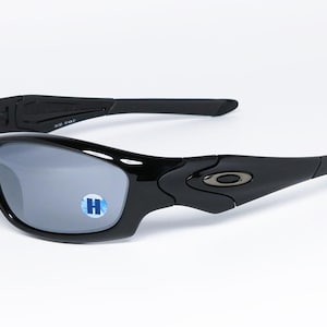 May include: Black sunglasses with gray lenses. The sunglasses have a curved frame and arms, with a small logo on the side. The lenses have a reflective quality. The sunglasses are on a white background.