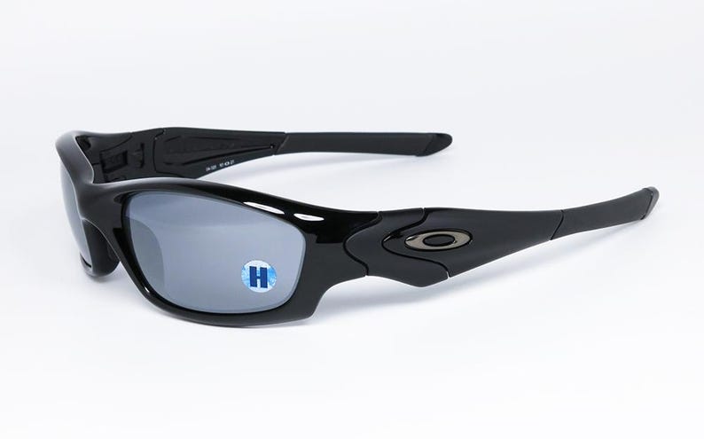 May include: Black sunglasses with gray lenses. The sunglasses have a curved frame and arms, with a small logo on the side. The lenses have a reflective quality. The sunglasses are on a white background.