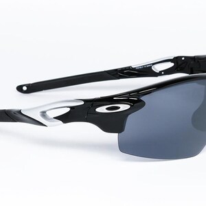 May include: Black and silver sports sunglasses with dark gray lenses. The sunglasses have a sleek, modern design with a curved frame and a prominent brand logo on the temples. The lenses are a dark gray color.